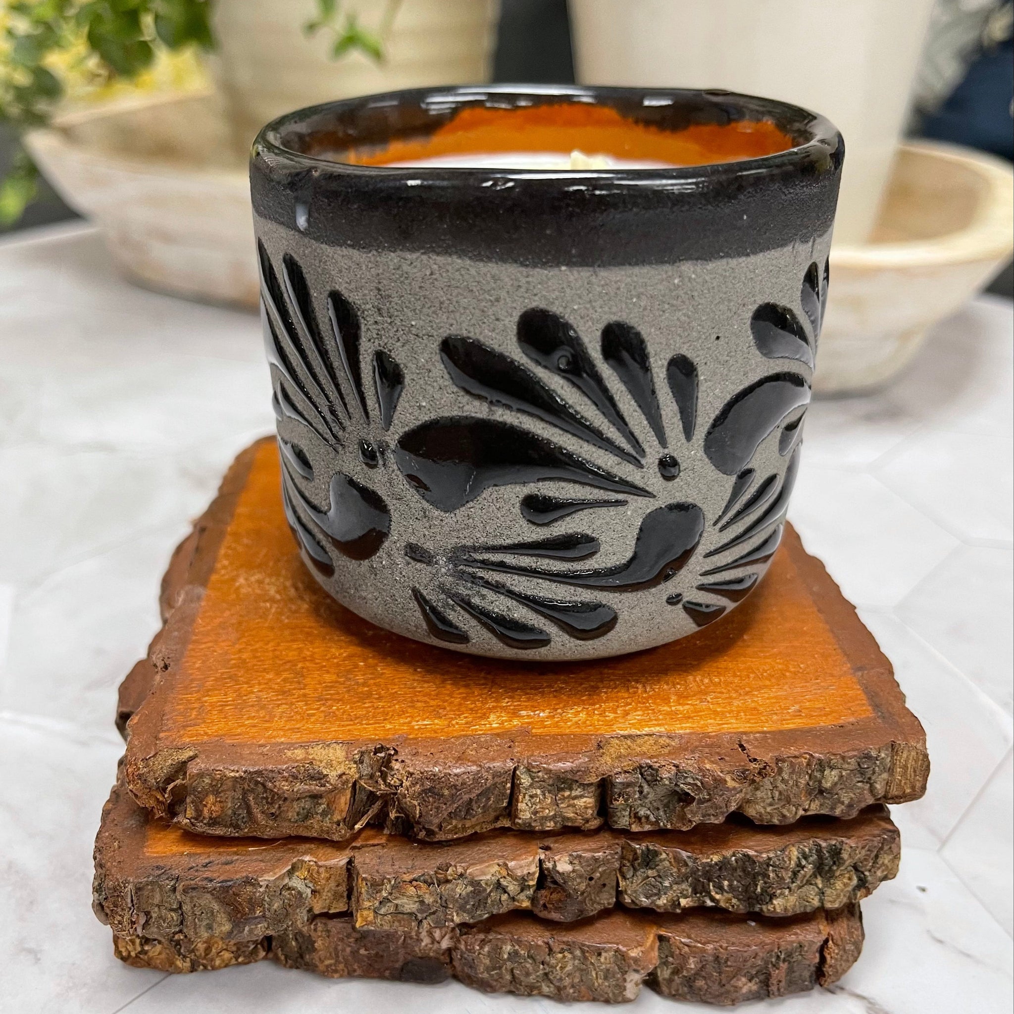 Handcrafted Soy Candle in Mexican black textured patterned Clay Vaso (6 oz) on top of 3 wooden coasters. potted plants are in the background.