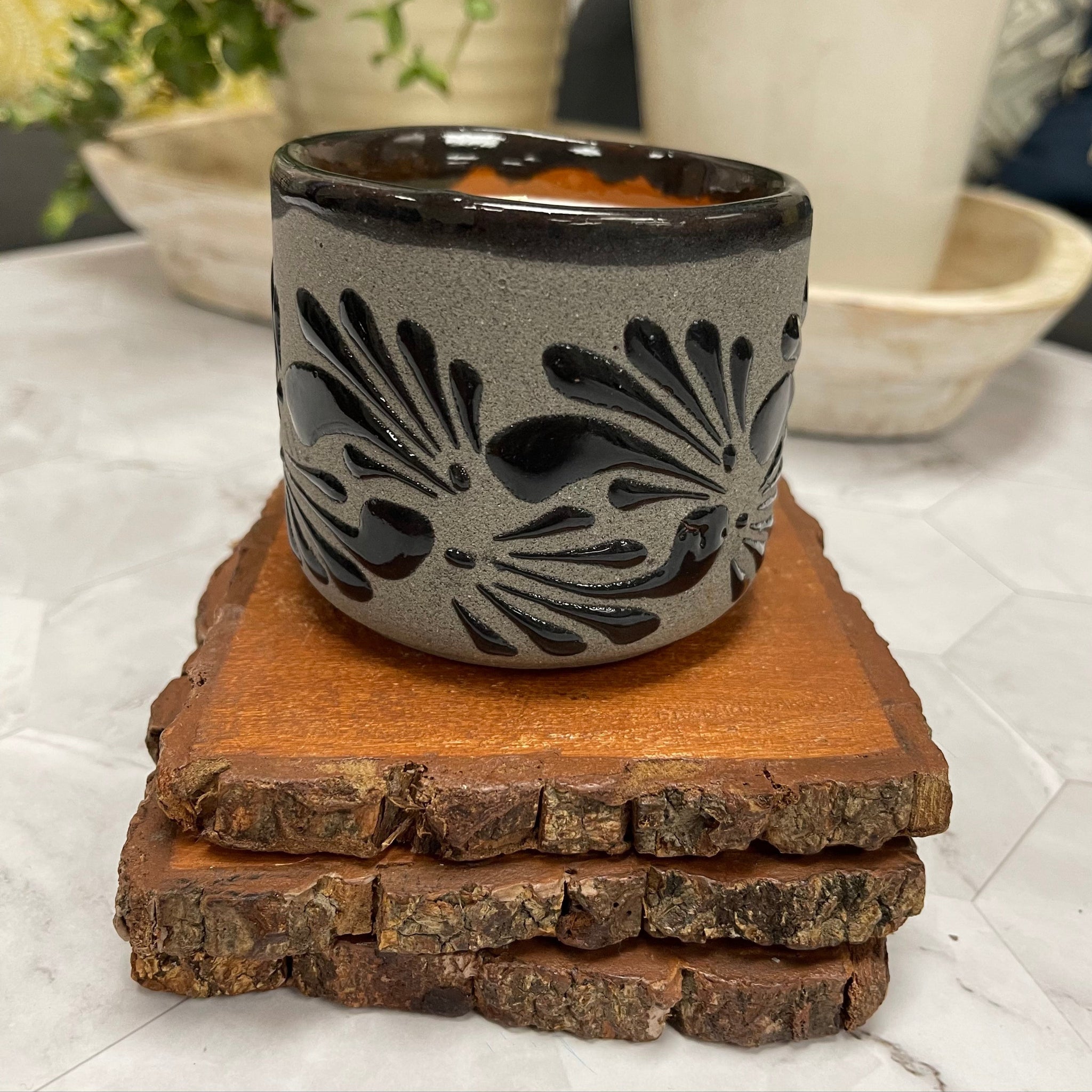 Handcrafted Soy Candle in Mexican black textured patterned Clay Vaso (6 oz) on top of 3 wooden coasters. potted plants are in the background.