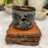 Handcrafted Soy Candle in Mexican black textured patterned Clay Vaso (6 oz) on top of 3 wooden coasters. potted plants are in the background.