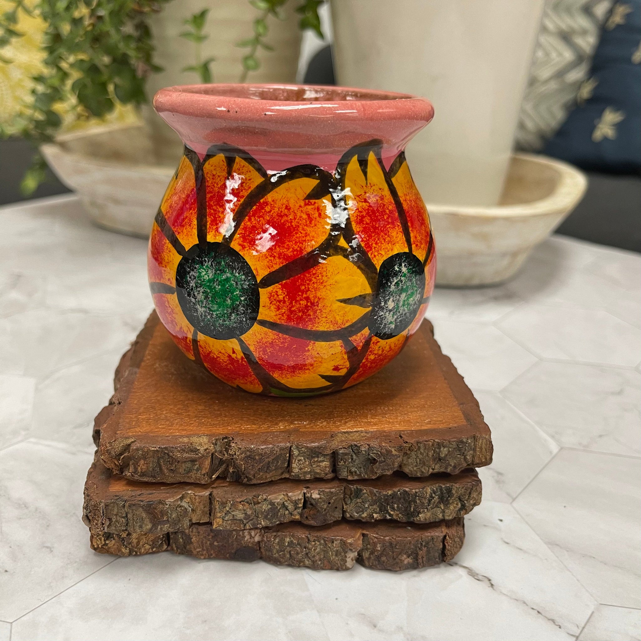 Floral handcrafted soy candle in Mexican clay Tacita in floral design on top of 3 wooden coasters. In the background there are 2 potted plants.