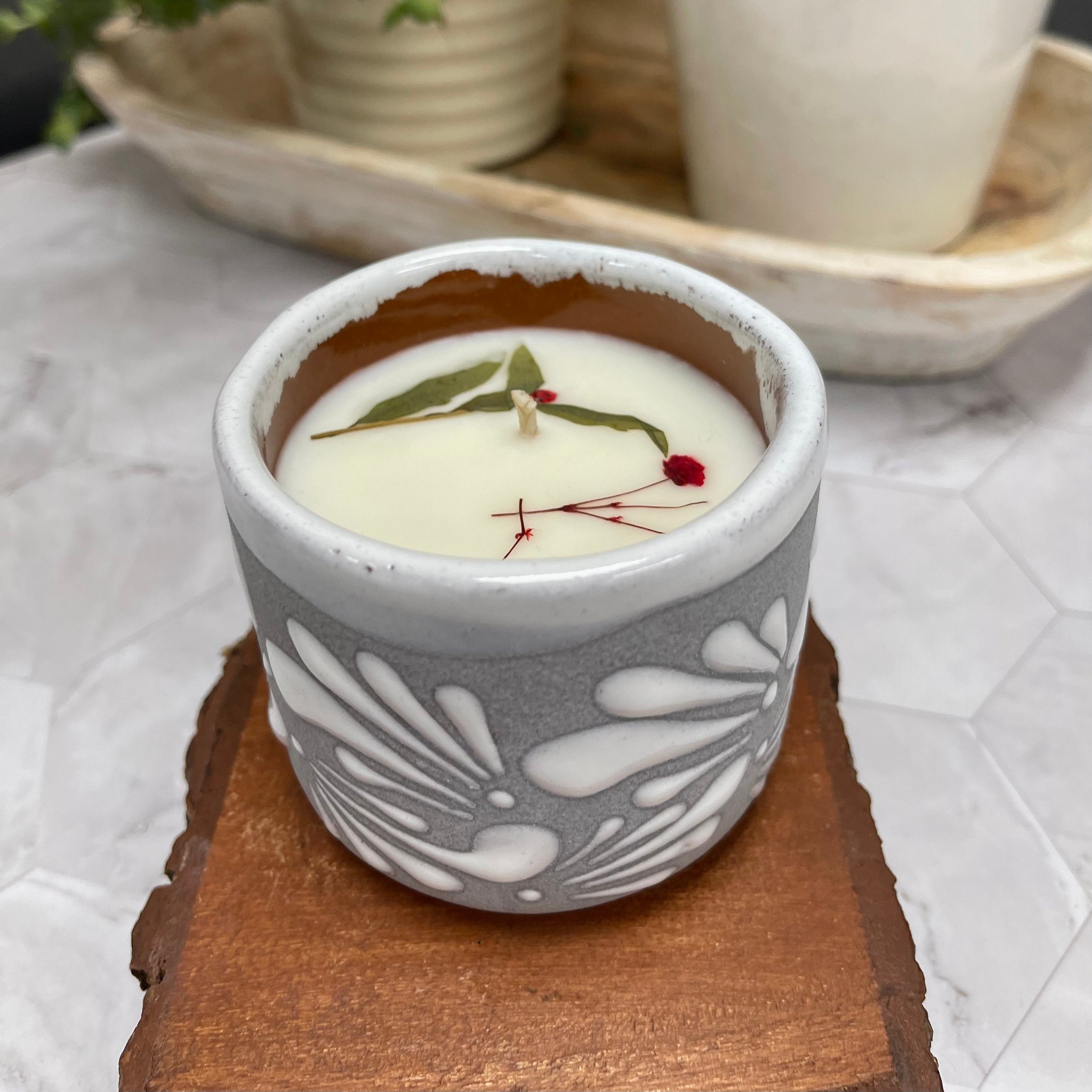 6 oz Mexican Clay Vaso candle in gray color sitting on top of wooden coasters. In the background there are potted plants.