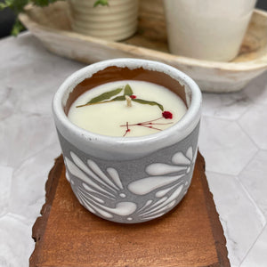6 oz Mexican Clay Vaso candle in gray color sitting on top of wooden coasters. In the background there are potted plants.