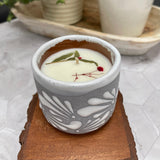 6 oz Mexican Clay Vaso candle in gray color sitting on top of wooden coasters. In the background there are potted plants.