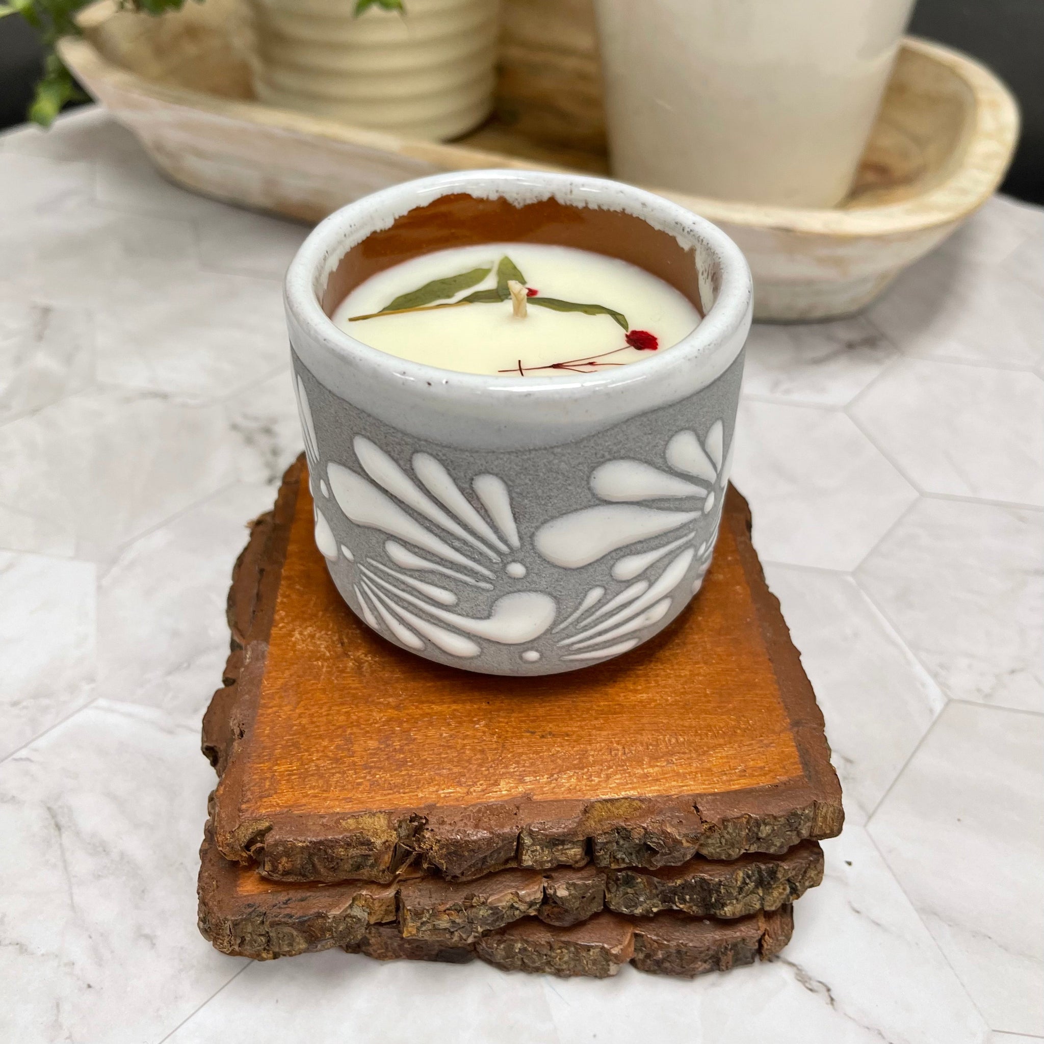 6 oz Mexican Clay Vaso candle in gray color sitting on top of wooden coasters. In the background there are potted plants.