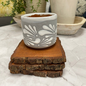 6 oz Mexican Clay Vaso candle in gray color sitting on top of wooden coasters. In the background there are potted plants.