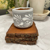 6 oz Mexican Clay Vaso candle in gray color sitting on top of wooden coasters. In the background there are potted plants.